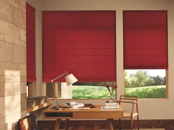 A cozy study room with wooden floors features a desk, a modern lamp, and a chair. Large windows adorned with custom blinds offer views of a lush green lawn outside, while red shades are partially drawn. A vase with flowers decorates the desk.