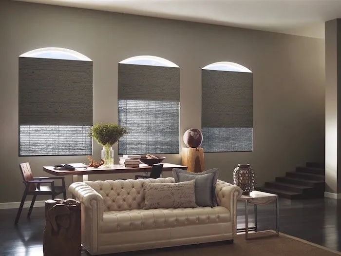 A modern living room features a white tufted sofa, a wooden desk with chairs, and lush greenery. Three tall, arched windows are adorned with elegant custom window treatments that complement the dark wood floor. A small staircase adds depth to the space.