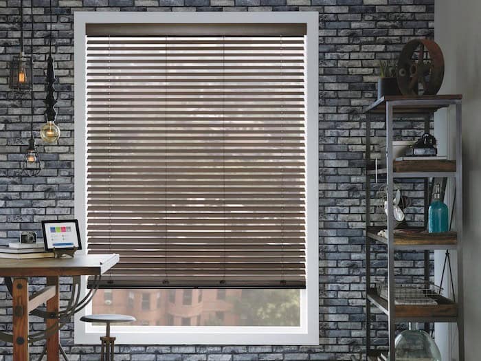 A room with a brick wall features a large window adorned with custom blinds. To the right, a metal shelf holds various industrial decorations. A small table on the left has a tablet and hanging light bulbs above, adding to the chic ambiance.