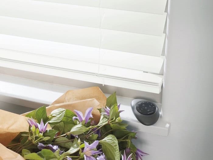 A bouquet of purple flowers with green leaves lies on a windowsill adorned with custom blinds. A small black circular remote rests beside the bouquet, and the blinds allow soft light to filter into the room.