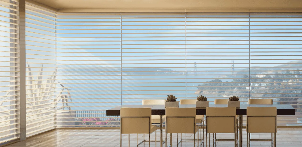 A dining room with a large table and white chairs is shown. Large windows with custom blinds offer a view of a bridge and body of water in the distance. Potted plants grace the table as natural light fills the room, creating a serene ambiance.