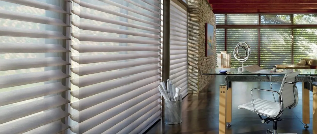 A modern office with floor-to-ceiling windows dressed in sleek window treatments. A glass desk with a white chair rests in the corner, accompanied by a round mirror and rolled-up papers nearby. The room's stone walls and wooden ceiling complete the sophisticated ambiance.