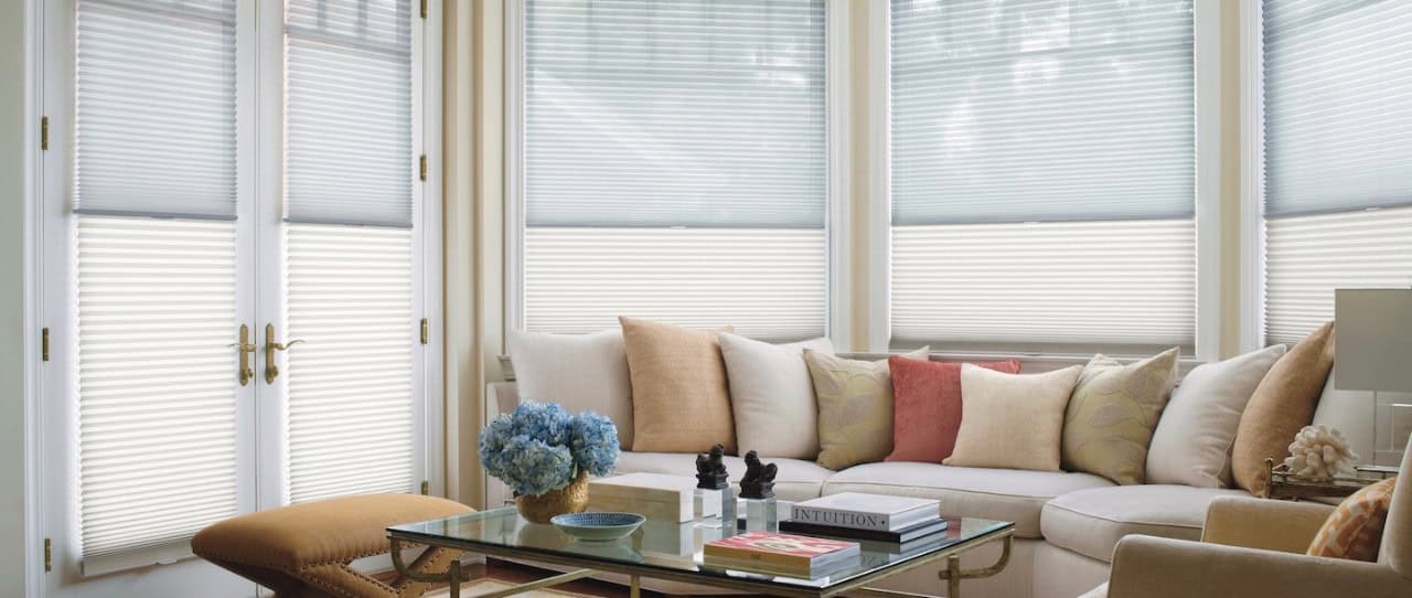 A cozy living room features a large beige sectional sofa adorned with various colored cushions. A glass coffee table holds books and a blue floral arrangement. Custom blinds cover multiple windows, perfectly complementing the orange ottoman in the foreground.