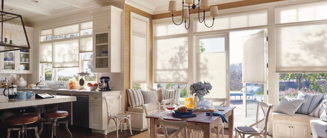 A bright kitchen and dining area with large windows adorned with beige Roman shades. The kitchen features a white counter with a tablet and a blue mixer. The dining table has chairs, flowers, and an outdoor view of the pool and greenery. A chandelier hangs above, casting elegance throughout.