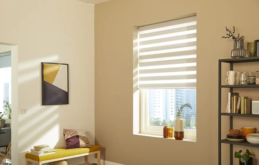 A cozy room with modern décor features custom blinds on the window, a yellow bench adorned with cushions and books, a geometric wall art piece, and a black shelving unit displaying dishes and decorative items. Sunlight filters through the blinds, casting gentle shadows.