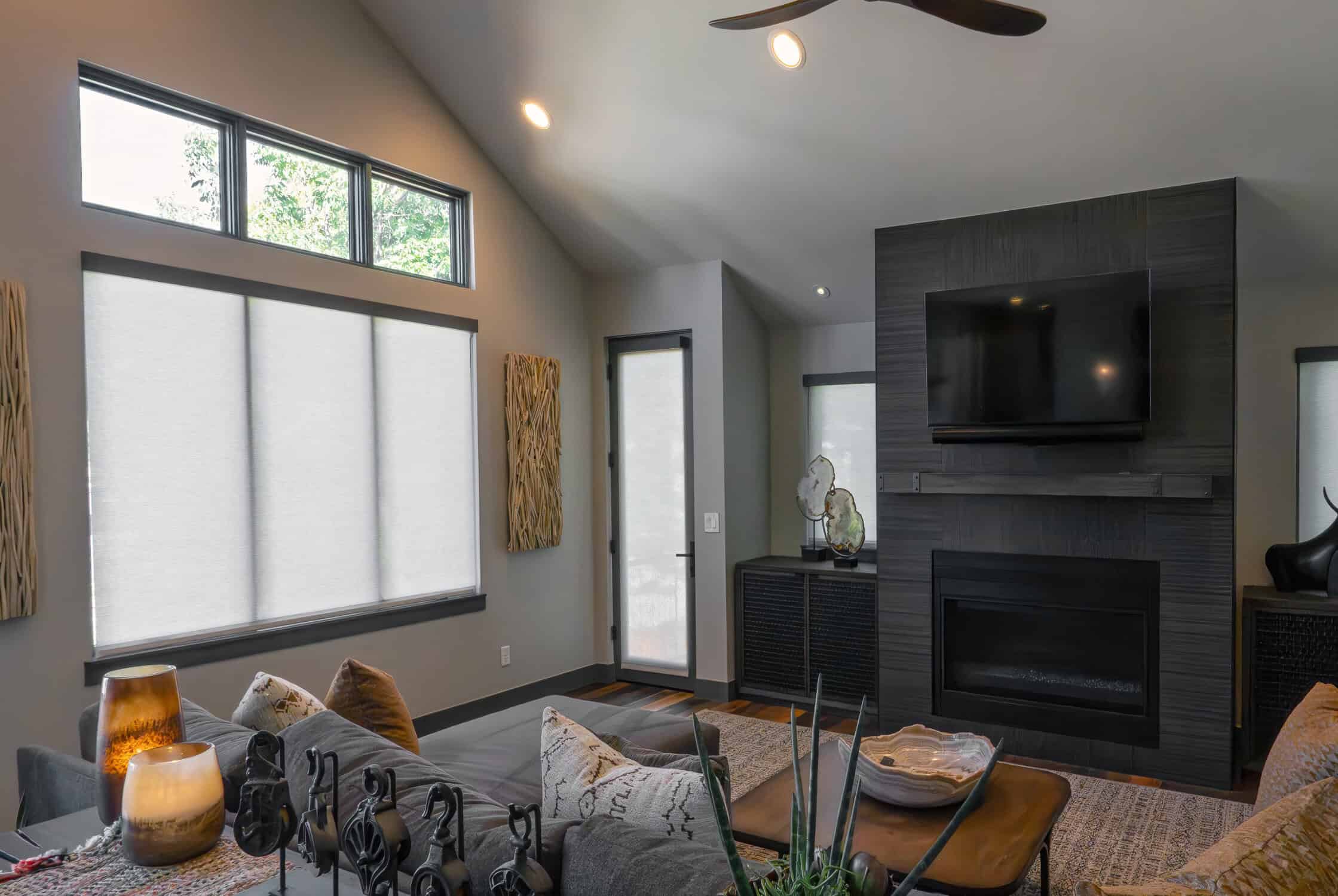 Modern living room with a dark gray accent wall featuring a flat-screen TV and fireplace. Large windows with sleek window shades let in natural light. Cozy seating, decorative cushions, candles, and sculptures add warmth to the contemporary space.