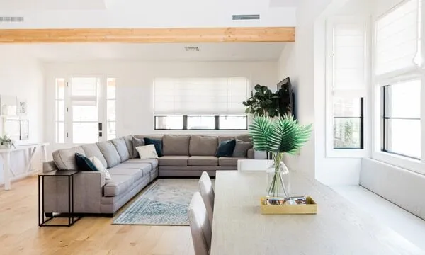 A bright living room featuring a large grey sectional sofa, a coffee table, and a flat-screen TV. In the foreground, a dining table boasts green fern decorations. Roman shades on the large windows allow natural light to flood the space, highlighting wooden beams and floor.
