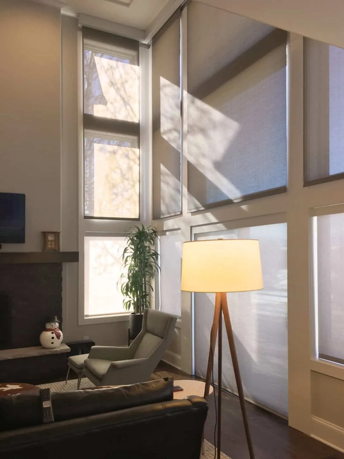 A modern living room with large floor-to-ceiling windows adorned with custom window treatments. Sunlight casts tree shadows on the shades. The room features a tall lamp, an armchair, a sofa, a fireplace, and a small snowman decoration.