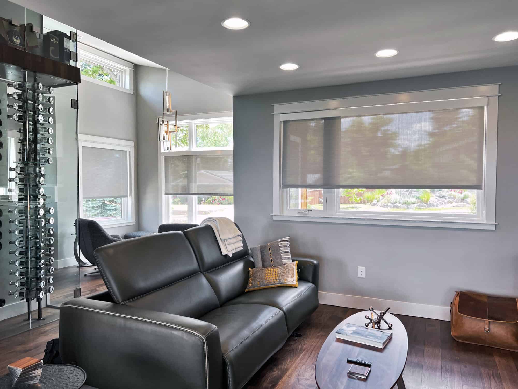 Modern living room with dark leather sofa, gray walls, and wooden flooring. Large windows showcase custom window treatments featuring sheer roller blinds. There's a unique glass and metal wine rack, an armchair, ceiling lights, and a wooden coffee table with magazines.