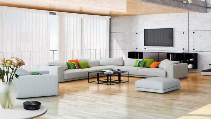 Spacious modern living room with light wood flooring, a large L-shaped gray sofa, colorful cushions, and a flat-screen TV on a concrete wall. Two black coffee tables stand near vertical custom window treatments, while a vase of roses adorns the side table.