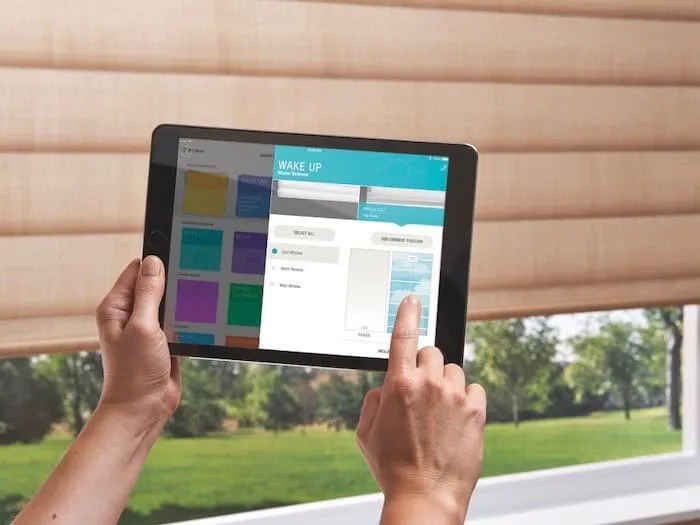 A person holds a tablet displaying a smart home app interface labeled "WAKE UP," featuring options for controlling custom window treatments. The background reveals partially drawn blinds, offering a glimpse of a lush garden and a sunny sky.