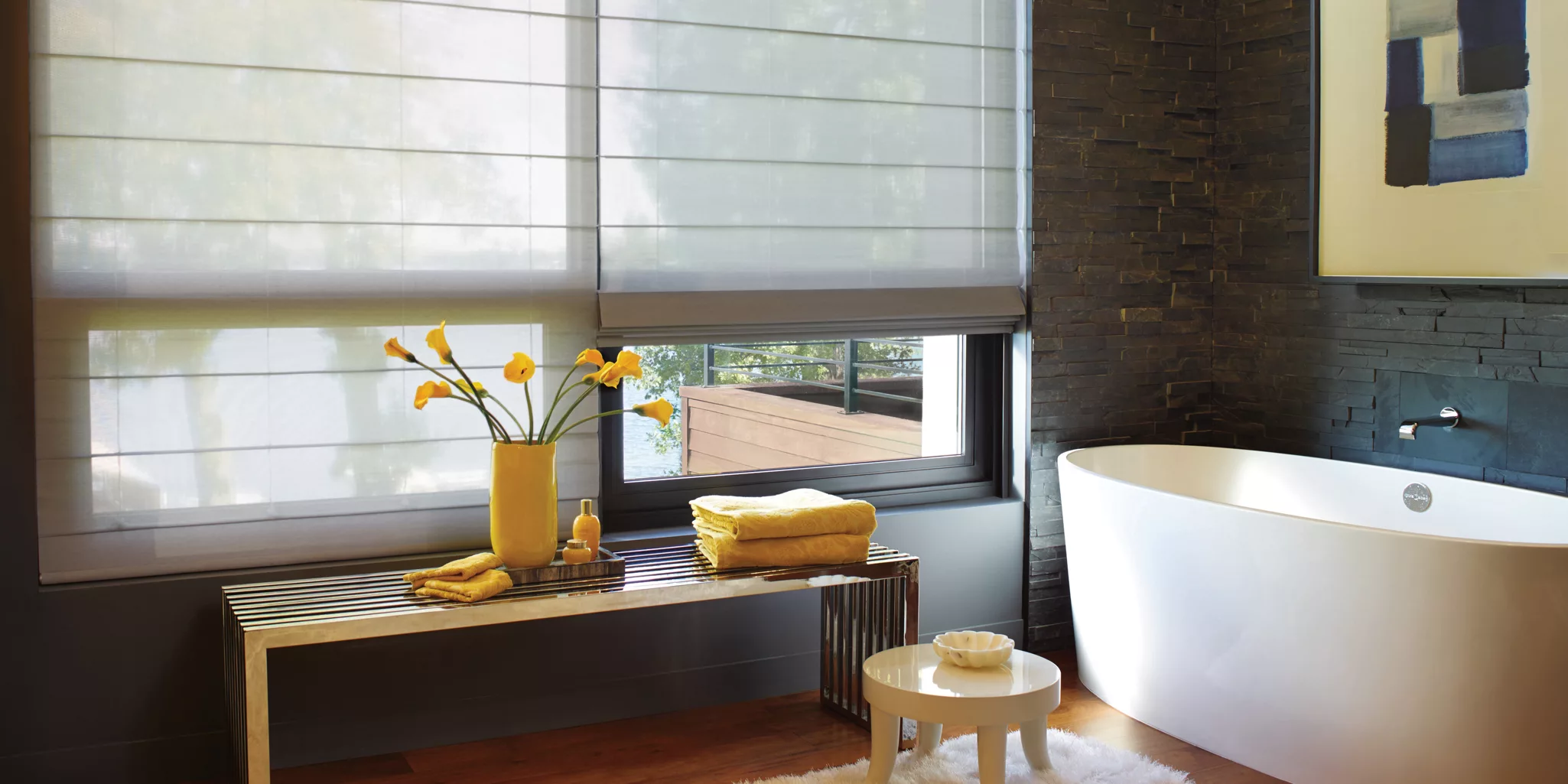 Modern bathroom with a freestanding white bathtub against a dark stone wall. A window with custom blinds softly filters the natural light. A wooden bench with a vase of yellow flowers and towels sits in the foreground, next to a small round table.