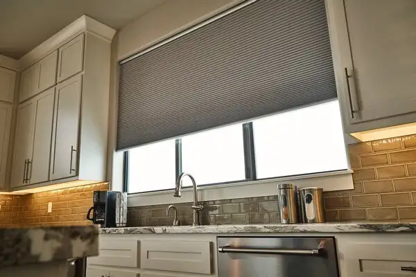 Kitchen interior with white cabinets and a marble countertop. A large window above the sink, adorned with custom blinds, features a gray pleated design partially lowered. The backsplash boasts beige subway tiles, and below sits a sleek stainless steel dishwasher.