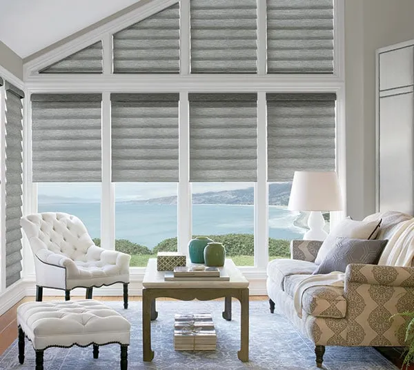 A bright living room with large windows featuring elegant gray window treatments. The space includes a patterned armchair, a tufted chair, and a wooden coffee table with vases and a table lamp. The windows offer a breathtaking view of the ocean and coastline.