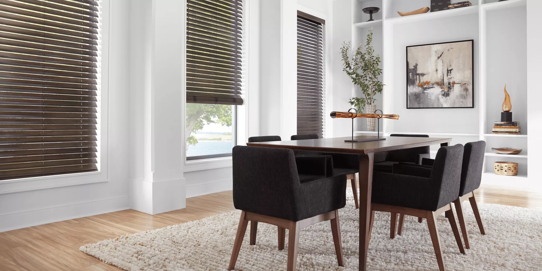 A modern dining room features a dark wooden table with six black upholstered chairs on a light textured rug. Large windows with custom blinds let in natural light, while a shelf with decor items and a framed abstract painting adorn the white walls.