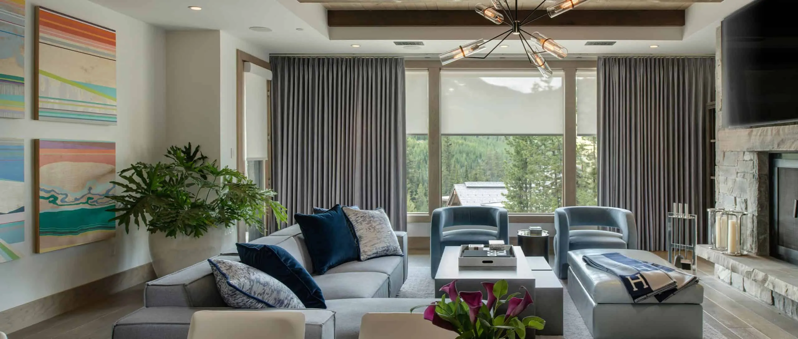 A modern living room with a gray sectional sofa, blue and white pillows, and blue armchairs is enhanced by custom window treatments framing the large window that showcases a scenic outdoor view. Abstract art adorns the wall, complemented by a stone fireplace and geometric light fixture.