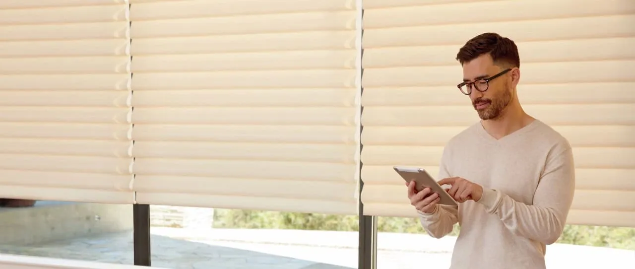 A man with a beard and glasses stands indoors using a tablet. He is wearing a light sweater. Behind him, large windows are adorned with elegant Roman Shades, allowing soft natural light to filter through the beige fabric.