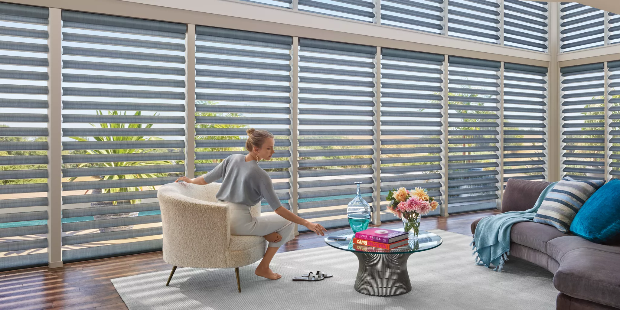 A person sits in a modern living room with large, sheer window shades. They reach for a cup on a stylish glass coffee table adorned with books, a flower vase, and a bottle. Nearby, a sofa with cushions rests on a light rug.