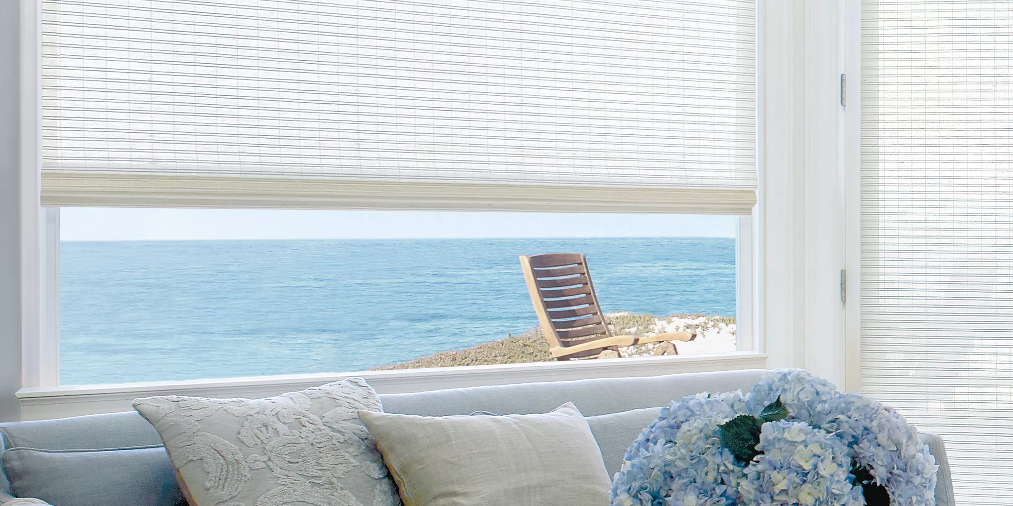 A sunlit room with a light gray couch and a bouquet of blue hydrangeas is adorned with elegant Roman shades. A window treatments enhance the view of the ocean, with a wooden chair facing the water, blending indoor comfort with a serene seaside landscape.