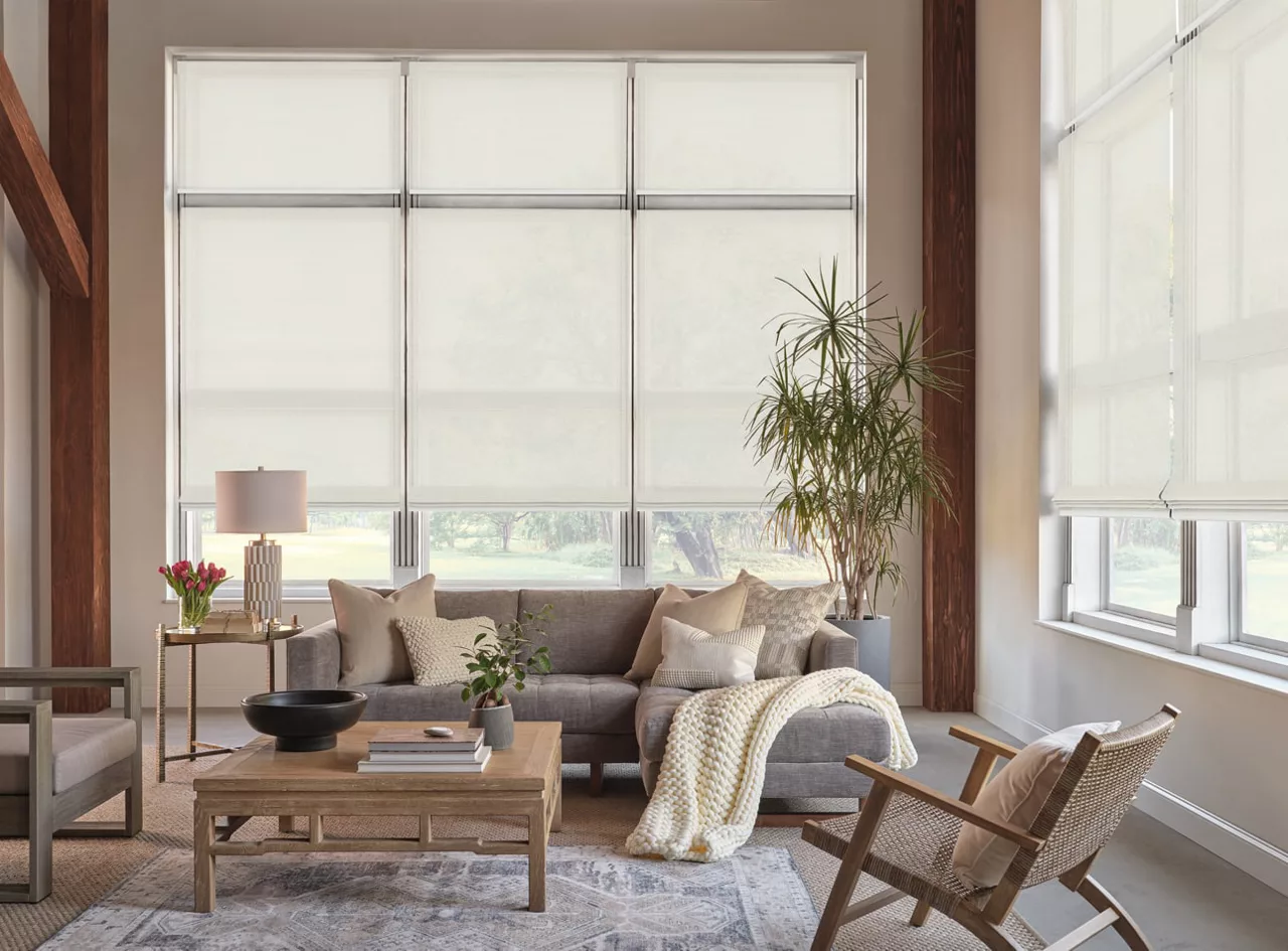 A modern living room with large windows covered by custom window shades. It features a gray sofa with pillows, a wooden coffee table adorned with decor, an armchair, a potted plant, and a side table with a lamp and flowers. Sunlight filters softly through the shades.