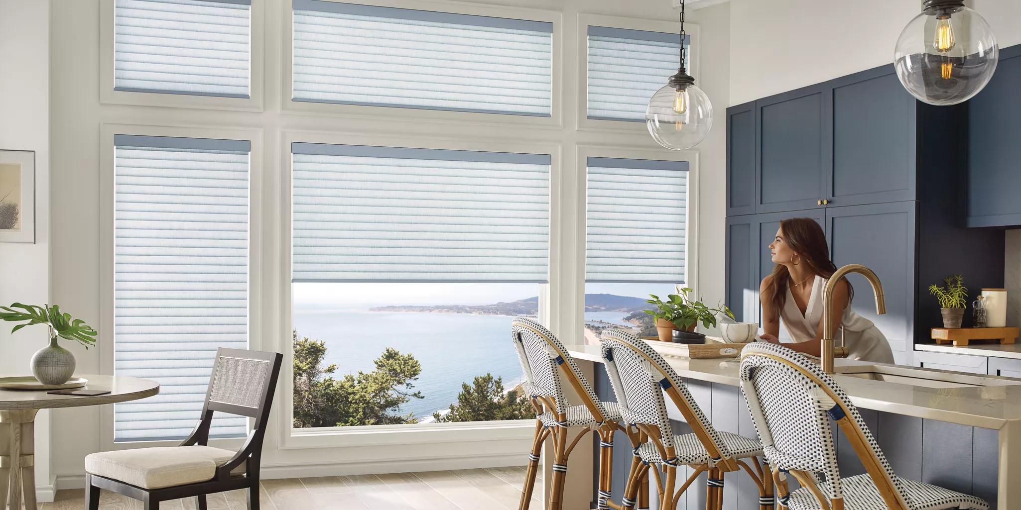 A woman stands in a stylish kitchen with blue cabinets and pendant lights. She gazes through large windows adorned with custom Roman shades, offering a scenic view of the ocean and coastline. Three stools line the kitchen island, and a small plant sits on a table.