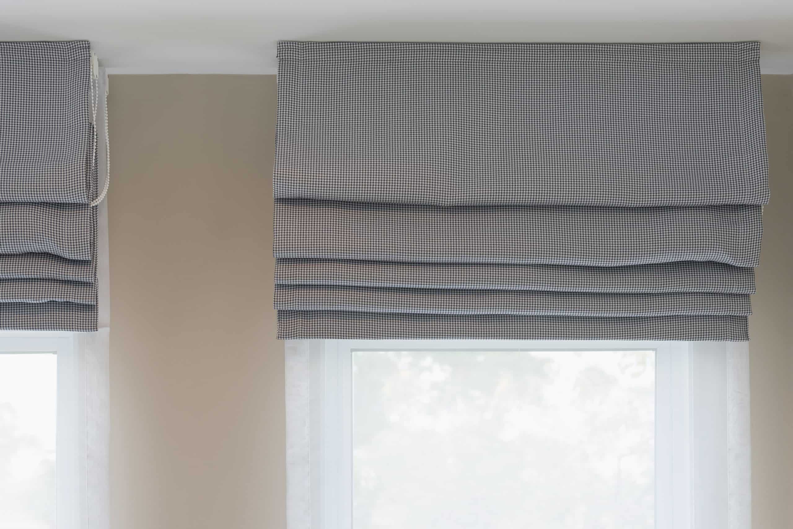Two windows with Roman shades partially drawn up. The shades feature a subtle checked pattern in grey and white, complementing the neutral beige wall color. Soft, natural light filters through the windows, creating a calm ambiance.