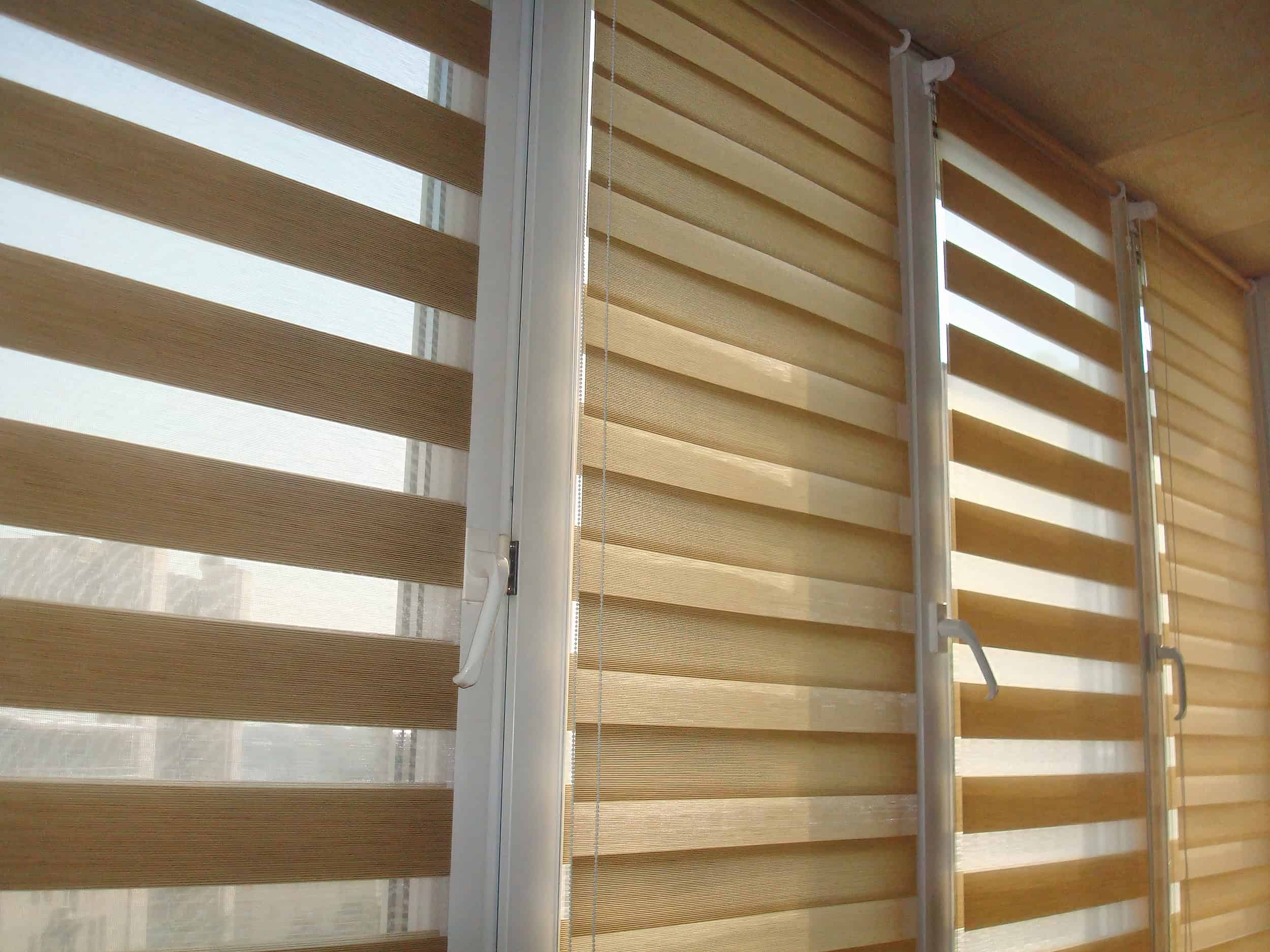 Vertical blinds with alternating sheer and opaque stripes cover a series of tall windows. Sunlight filters through, creating soft shadows on the floor. The view outside shows a blurred cityscape in the background.