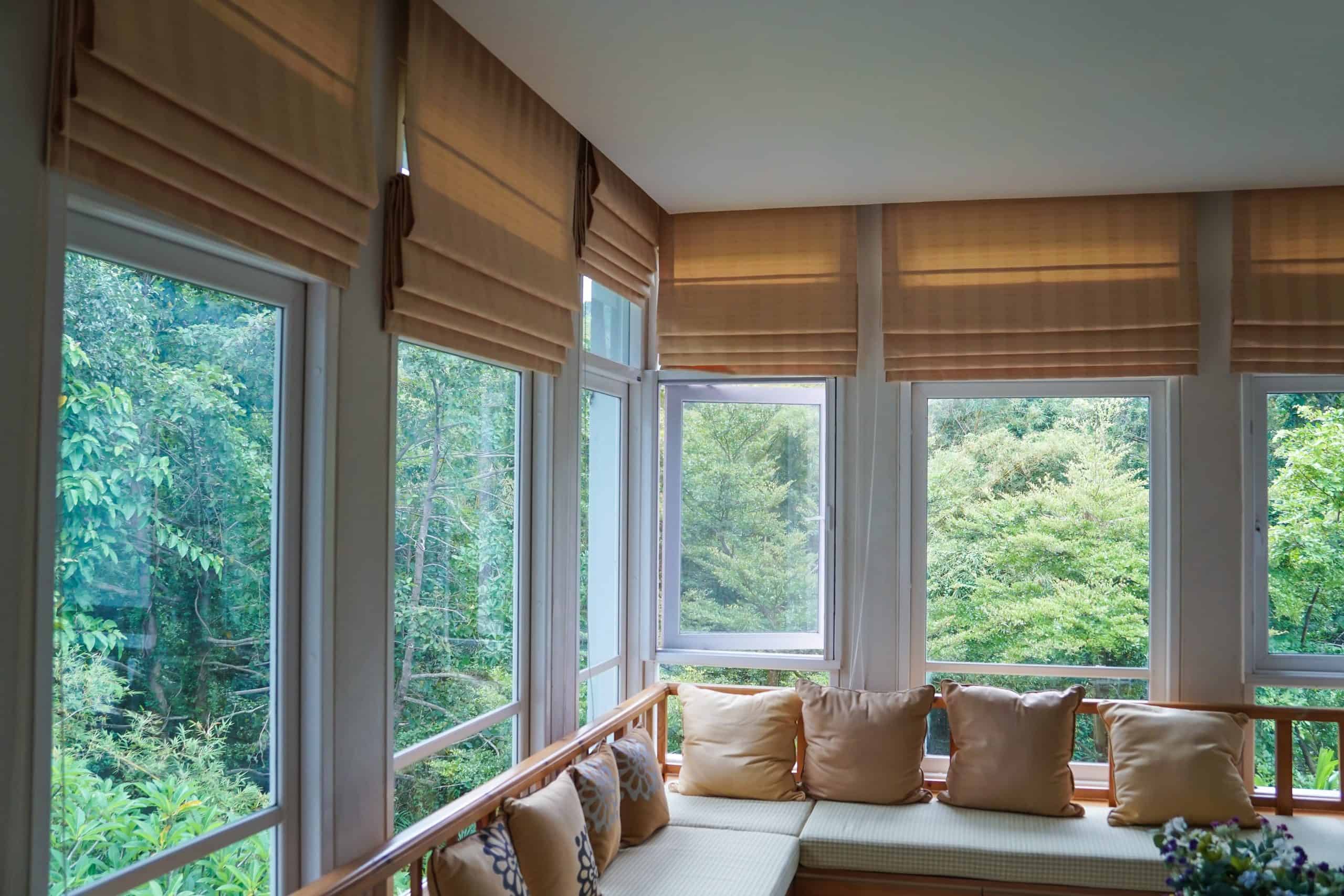 A cozy sunroom with large windows and beige Roman shades. The seating area features a wooden bench lined with beige cushions, offering views of lush green trees outside. Natural light fills the space, creating a serene atmosphere.