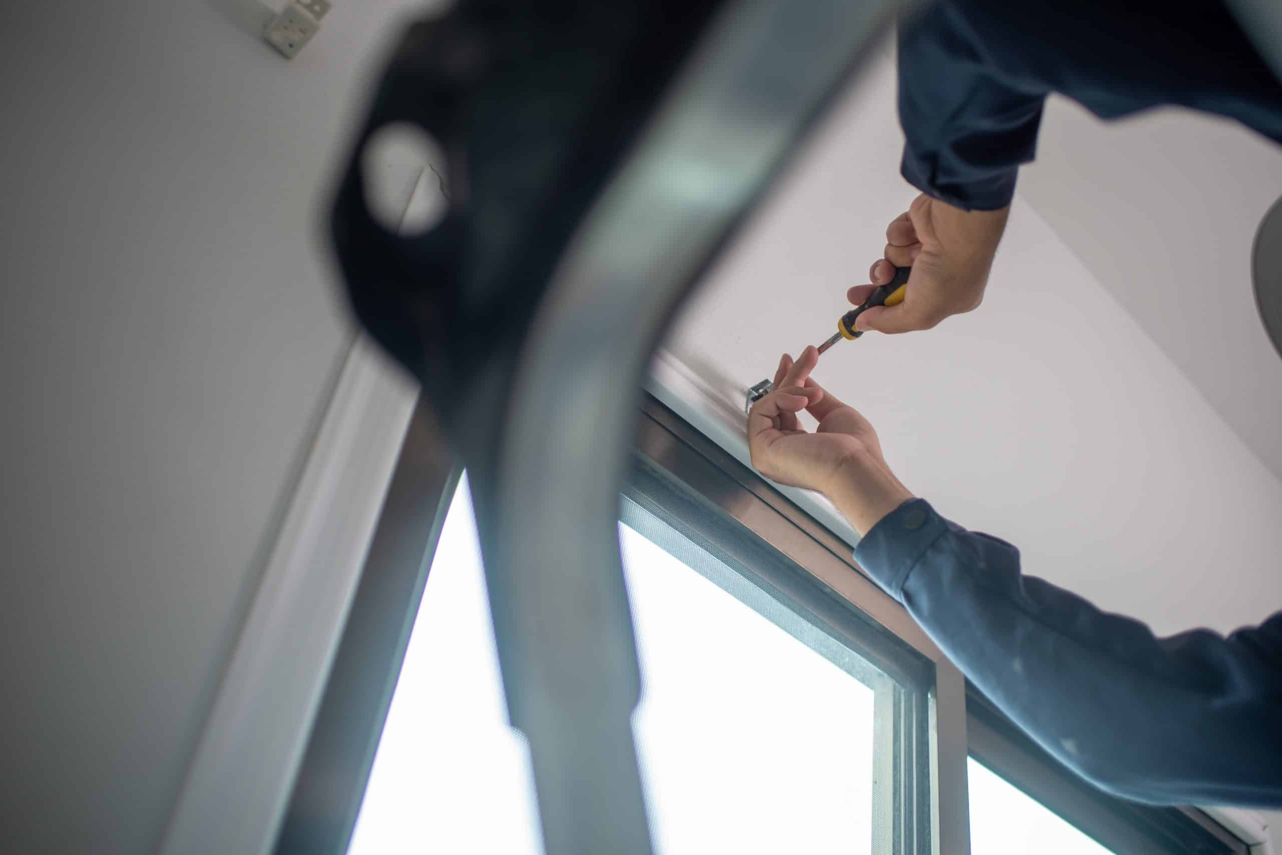 Person in work attire using a screwdriver to adjust a window or frame from a ladder's perspective. The focus is on their hands and tool, with part of the window and ladder visible in the foreground.