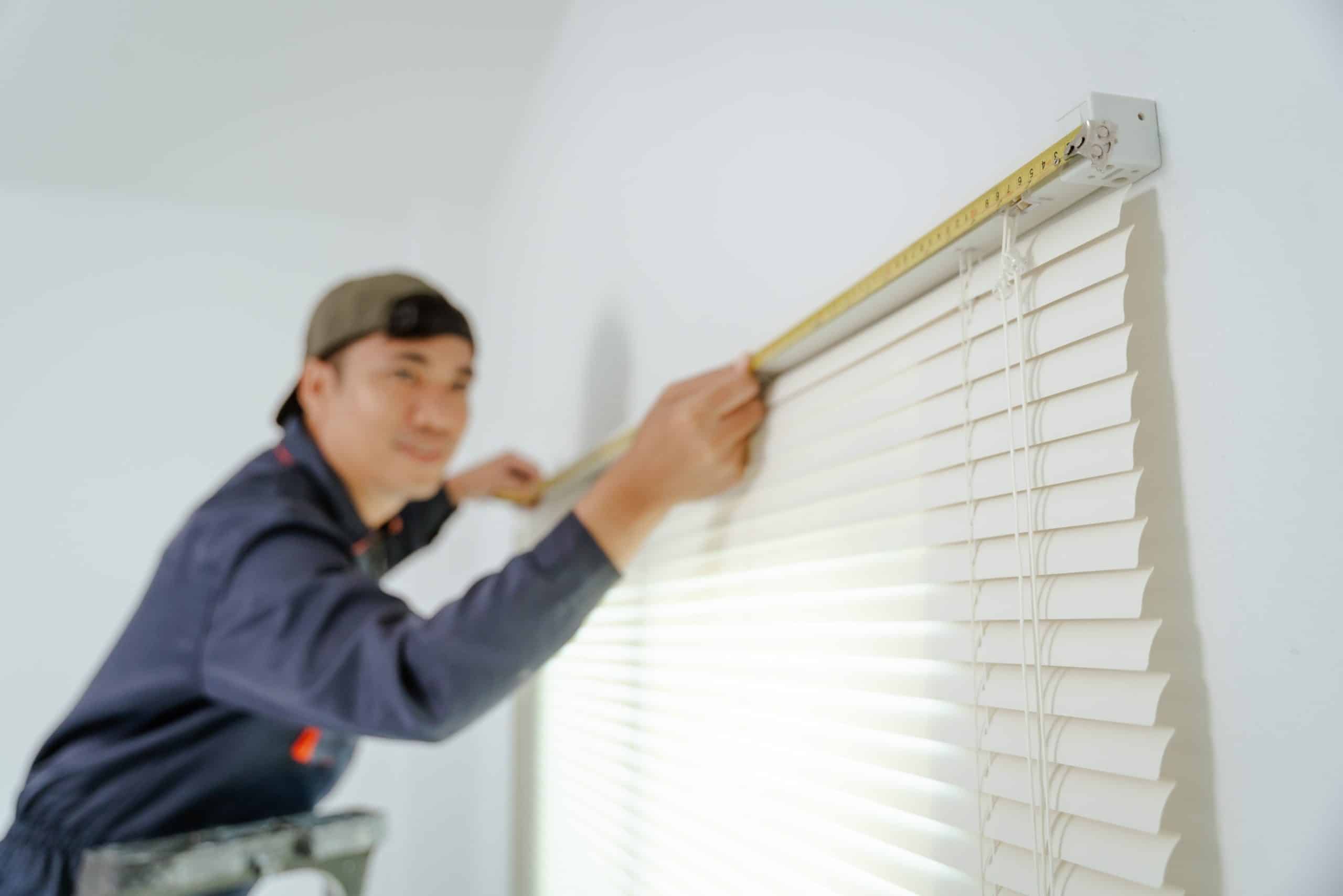 A person in a cap and work uniform measures a white window blind with a tape measure. They are focused on the task, standing on a ladder against a white wall. The room is brightly lit.