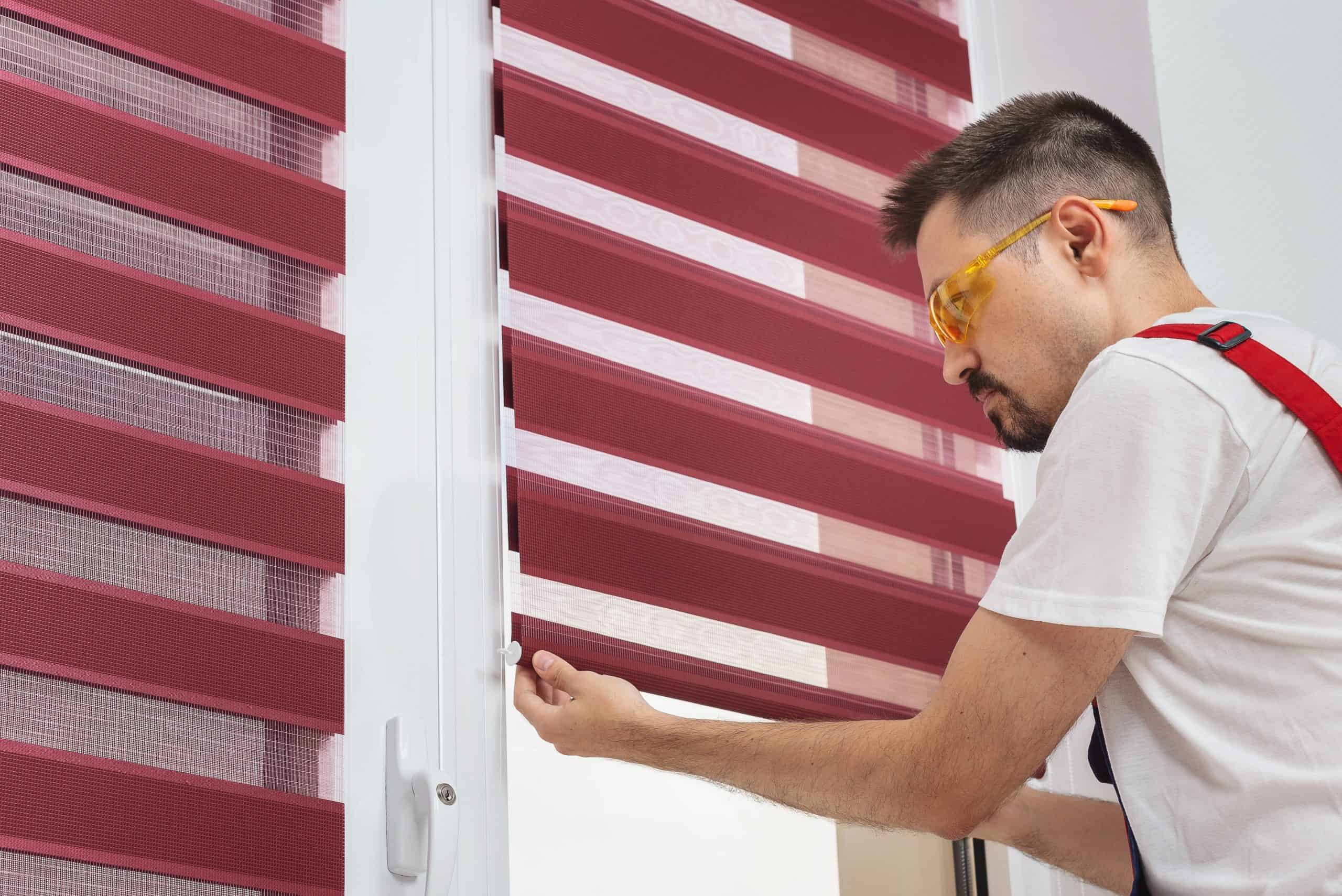 A person wearing yellow safety glasses and a white shirt adjusts a modern red roller blind on a window. The blinds consist of alternating sheer and opaque horizontal stripes, allowing varying light control.