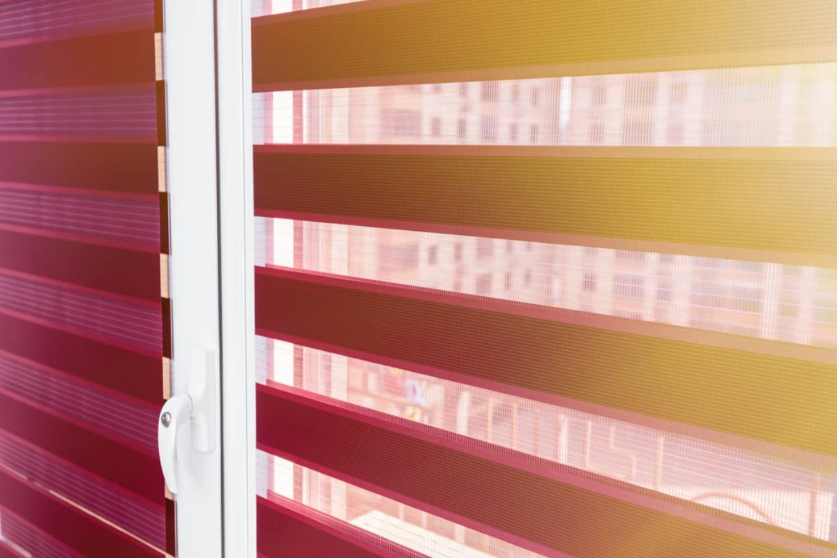 Close-up view of a white-framed window with red and translucent zebra blinds, allowing filtered light to enter. The blurred cityscape is visible through the blinds.
