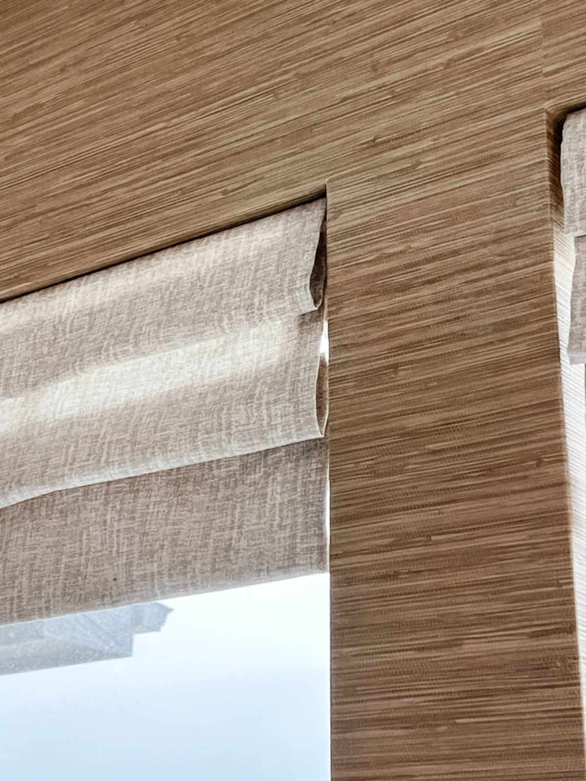 Close-up of a window with beige textured Roman shades partially raised, letting in natural light. The wall and window frame have a matching light wood grain pattern.