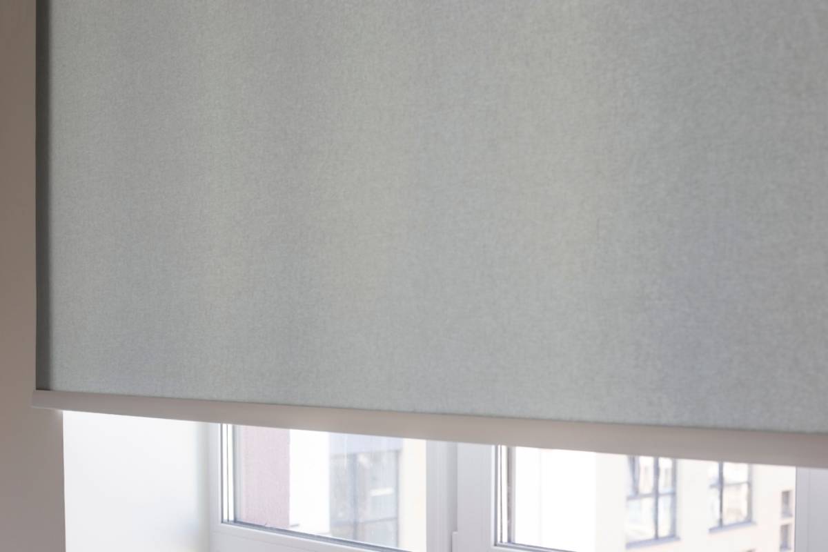 A close-up of a window with a light gray roller blind partially lowered, allowing some natural light to filter through. Parts of window frames and building exteriors are faintly visible below the blind.