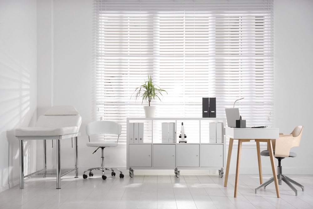 A modern, minimalistic medical office with white furniture, an exam table, swivel chairs, a desk with a laptop, cabinets, folders, and a potted plant in front of a large window with white blinds.