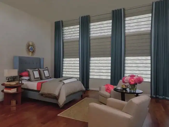 A bedroom with a bed with a gray headboard, and a set of three large windows covered by light-filtering horizontal shades and dark blue drapery panels. Two armchairs and a small round table with a bouquet of pink flowers are in front of the windows.
