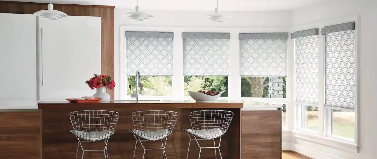 A modern kitchen with a large window over the sink, covered by three light-colored roller shades with a subtle geometric pattern. A kitchen island with three wire-frame bar stools is in the foreground.