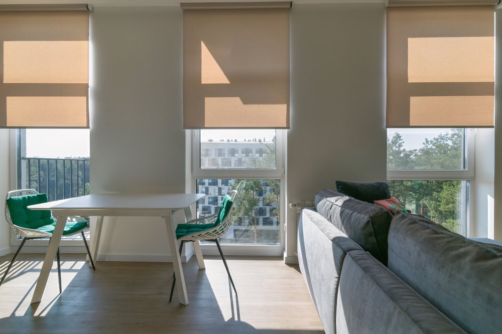 A modern living room with large windows covered by beige blinds, a white table with two green chairs, and a gray sofa. Sunlight filters through the windows, creating soft shadows on the wooden floor.
