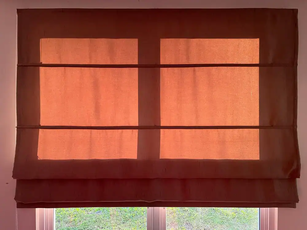 A close-up shot of a warm, orange fabric Roman blind partially covering a window. The sunlight filtering through the fabric creates a glowing effect, highlighting the horizontal folds of the blind.