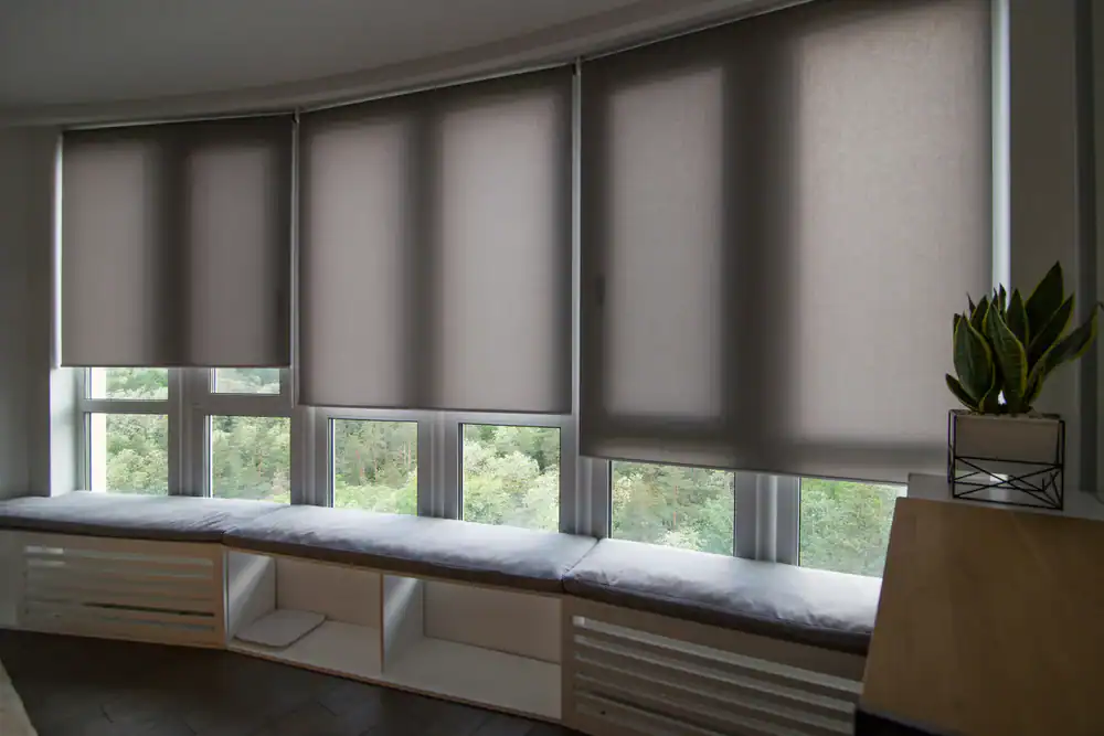 A modern living space with a large, curved bay window. The window is fitted with gray roller blinds and has a long, cushioned window seat with storage underneath.